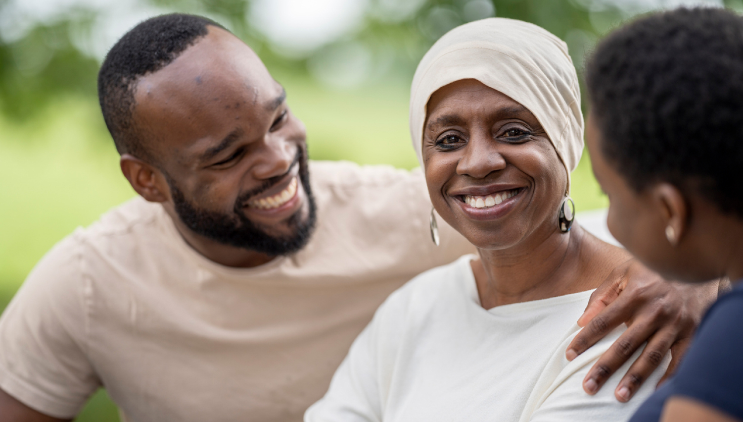 Smiling cancer survivor with headscarf surrounded by supportive family members during outdoor conversation, highlighting the emotional impact of Cancer Care Costs.